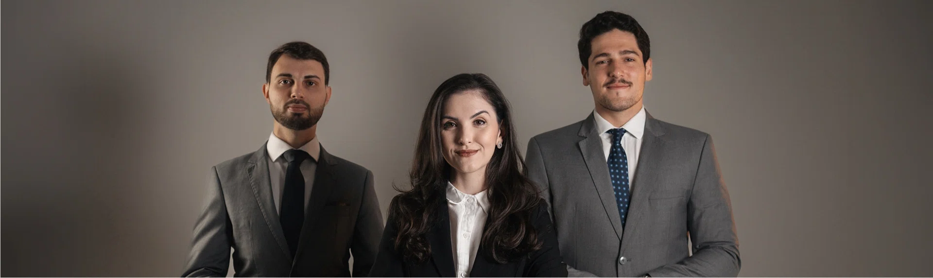 Time de advogados sócios: Maria, Arthur e Marcelo em trajes sociais contra fundo cinza.